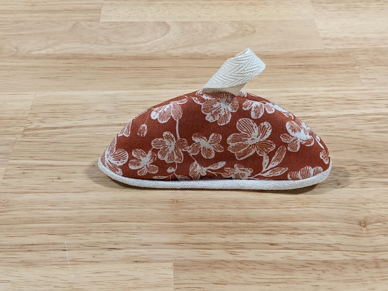 May include: A dome-shaped fabric pot handle cover featuring a rust-colored background with a white floral pattern. The item includes a white herringbone-patterned fabric loop at the top and is finished with a white fabric trim along the bottom edge.