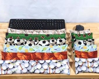 May include: A collection of rectangular fabric wrist rests featuring various sports-themed patterns including baseballs, footballs, soccer balls, pickleball paddles, basketballs, and golf balls with tees, arranged in front of a black computer keyboard.