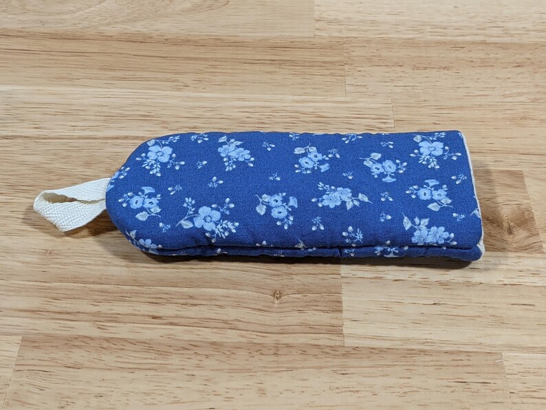 May include: A rectangular quilted oven mitt in a vibrant blue fabric featuring a repeating pattern of small light blue flowers. The mitt includes a white fabric loop attached to one end for hanging, providing a functional design for kitchen use.