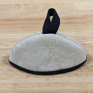 May include: A semi-circular, light gray fabric cover with a textured weave. The item features a dark navy blue fabric trim along the bottom edge and a matching dark navy blue fabric loop handle attached to the top center for easy lifting.