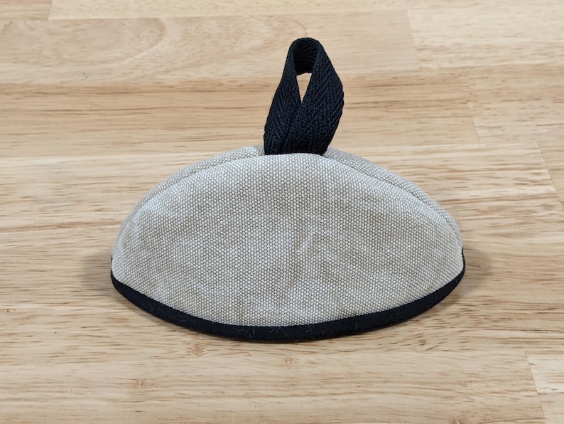 May include: A semi-circular, light gray fabric cover with a textured weave. The item features a dark navy blue fabric trim along the bottom edge and a matching dark navy blue fabric loop handle attached to the top center for easy lifting.