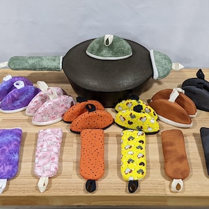 May include: A black cast iron pot with a green textured lid cover and handle covers, surrounded by various fabric pot holders and handle covers in purple, pink, orange, yellow with chicken prints, brown, and black, all featuring hanging loops.