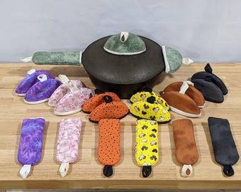 May include: A black cast iron pot with a green textured lid cover and handle covers, surrounded by various fabric pot holders and handle covers in purple, pink, orange, yellow with chicken prints, brown, and black, all featuring hanging loops.