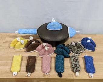 May include: A black cast iron skillet with a blue fabric handle cover and lid knob cover, surrounded by various fabric pot holders and handle covers in yellow, brown, pink, teal, camouflage, and blue patterns, each featuring a small white hanging loop.