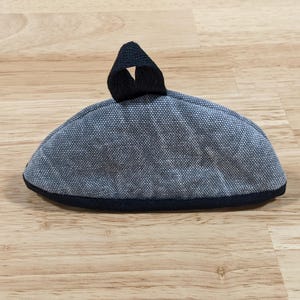 May include: A semi-circular fabric pot holder in a textured grey and white weave. The item features a solid black fabric trim along the bottom edge and a small black fabric loop attached to the top center for hanging or storage purposes.