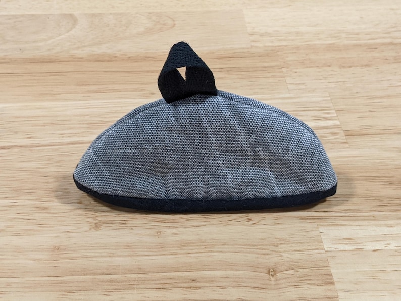 May include: A semi-circular fabric pot holder in a textured grey and white weave. The item features a solid black fabric trim along the bottom edge and a small black fabric loop attached to the top center for hanging or storage purposes.