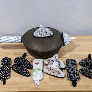 May include: A dark gray cast iron pot with matching cat-themed handle covers and lid. Several cat-themed oven mitts and pot holders in various designs, including black and white polka dots, and cat illustrations, are arranged around the pot.
