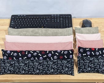 May include: A collection of rectangular fabric wrist rests and smaller matching cushions arranged in front of a black computer keyboard and mouse. The patterns include solid light gray, solid light pink, pink with white dots, black with colorful paw prints and bones, and black with white cat outlines.