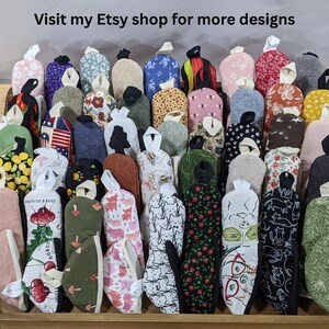 May include: A collection of small, fabric oven mitts in a variety of patterns and colors, including floral, animal, and geometric prints. Each mitt features a small hanging loop at the top. The text 'Visit my Etsy shop for more designs' is visible.