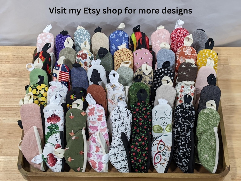 May include: A collection of small, fabric oven mitts in a variety of patterns and colors, including floral, animal, and geometric prints. Each mitt features a small hanging loop at the top. The text 'Visit my Etsy shop for more designs' is visible.