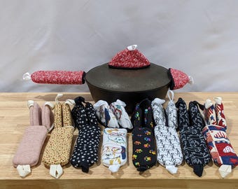 May include: A black cast iron pot with a red patterned lid cover and handle covers, surrounded by a collection of various fabric handle covers in assorted colors and patterns including floral, paw prints, dog faces, and patriotic heart designs.