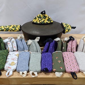 May include: A cast iron pot with a lemon-patterned lid cover and two matching handle covers, surrounded by a collection of rectangular and triangular fabric pot holders in various colors and patterns including bees, mushrooms, and solid textures.