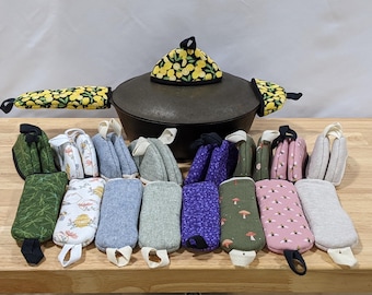 May include: A cast iron pot with a lemon-patterned lid cover and two matching handle covers, surrounded by a collection of rectangular and triangular fabric pot holders in various colors and patterns including bees, mushrooms, and solid textures.