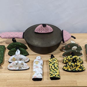 Op de afbeelding: Een verzameling keukenaccessoires, waaronder een donkere metalen pot met roze stippen handgreephoezen en deksel. Extra items zijn groene pannenlappen met bladeren, items met paddenstoelthema en accessoires met bijen- en citroenpatronen.