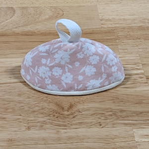 May include: A dome-shaped fabric tea cozy featuring a soft pink background with a white floral pattern. The item has a white fabric loop handle at the top center and a white fabric trim along the bottom edge, providing a clean and finished look.