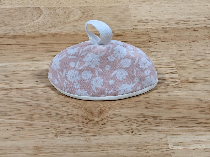 May include: A dome-shaped fabric tea cozy featuring a soft pink background with a white floral pattern. The item has a white fabric loop handle at the top center and a white fabric trim along the bottom edge, providing a clean and finished look.