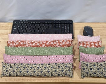 May include: A collection of five long rectangular fabric wrist rests and five matching smaller rectangular fabric wrist rests, each featuring distinct floral, botanical, and bee patterns in shades of pink, rust, green, and cream, arranged near a keyboard.