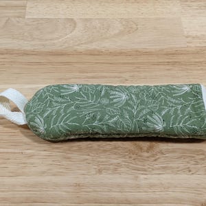 May include: A rectangular quilted fabric handle cover in sage green featuring a white botanical print of leaves and stems. The cover includes a white fabric loop on one end and white trim along the edges, designed for a kitchen pan handle.