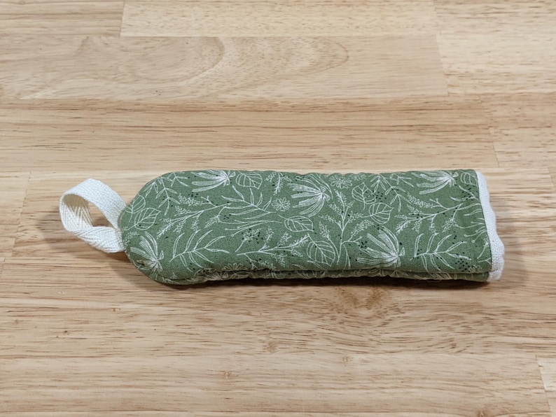 May include: A rectangular quilted fabric handle cover in sage green featuring a white botanical print of leaves and stems. The cover includes a white fabric loop on one end and white trim along the edges, designed for a kitchen pan handle.