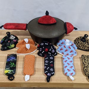 May include: A collection of fabric kitchen accessories including a dark cast iron pot with a red velvet lid cover and handle covers, alongside various patterned pot holders and handle sleeves featuring vegetable, polka dot, paw print, lobster, and leopard prints.