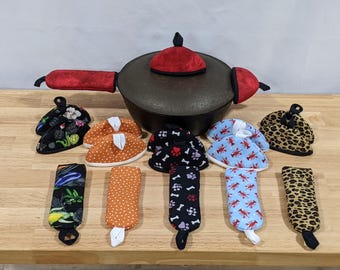 May include: A collection of fabric kitchen accessories including a dark cast iron pot with a red velvet lid cover and handle covers, alongside various patterned pot holders and handle sleeves featuring vegetable, polka dot, paw print, lobster, and leopard prints.