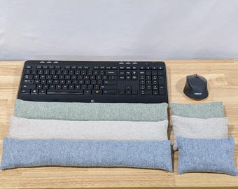 May include: A black computer keyboard and mouse positioned above three long, rectangular fabric wrist rests in sage green, light gray, and heather blue, accompanied by three matching small rectangular fabric cushions to the right side.