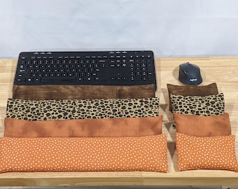 May include: A black computer keyboard and mouse positioned above four rectangular fabric wrist rests. The rests feature four distinct patterns: a dark brown textured fabric, a leopard print, a solid burnt orange, and an orange with white polka dots.