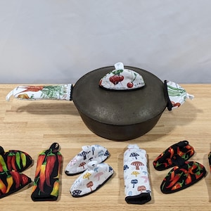 May include: A dark grey cast iron pot with fabric handle covers and a lid with a fabric knob. Several matching pot holders and handle covers are arranged around the pot. The fabric patterns include vegetables, mushrooms, and chilli peppers.