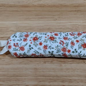 May include: A rectangular fabric pouch featuring a white background with a repeating floral pattern of small orange flowers and green leaves. The pouch has a rounded end and a small white fabric loop attached to one side for hanging or carrying.