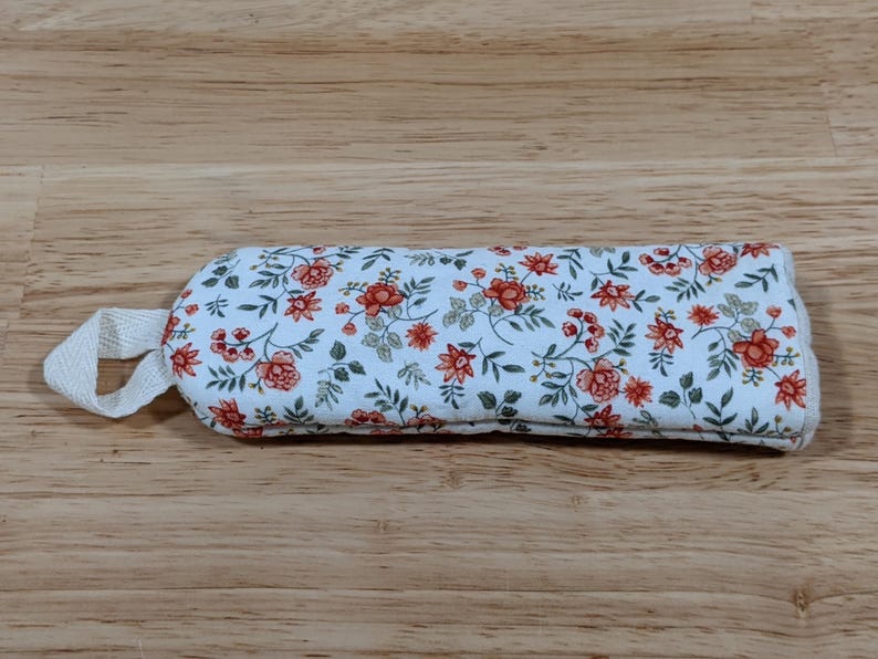 May include: A rectangular fabric pouch featuring a white background with a repeating floral pattern of small orange flowers and green leaves. The pouch has a rounded end and a small white fabric loop attached to one side for hanging or carrying.