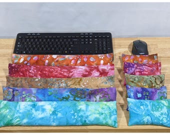 May include: A collection of rectangular fabric wrist rests in various tie-dye patterns, including orange, red, brown, purple, and blue. These soft cushions are arranged in a stack below a black computer keyboard and next to a black computer mouse.
