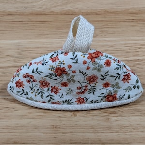 May include: A semi-circular fabric pot handle cover featuring a white background with a repeating floral pattern of orange flowers and green leaves. The item includes a beige fabric loop at the top and a beige fabric trim along the bottom edge.