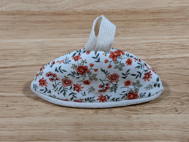 May include: A semi-circular fabric pot handle cover featuring a white background with a repeating floral pattern of orange flowers and green leaves. The item includes a beige fabric loop at the top and a beige fabric trim along the bottom edge.