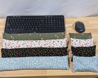 May include: A collection of four long rectangular fabric wrist rests and four matching small rectangular fabric cushions. The patterns include an olive green with mushrooms, a white floral print, a black rose print, and a light blue leaf pattern.