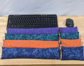 May include: A collection of four long rectangular fabric wrist rests and four matching small rectangular fabric cushions. The items feature purple, teal, orange with black dots, and blue marbled patterns, arranged near a black computer keyboard.