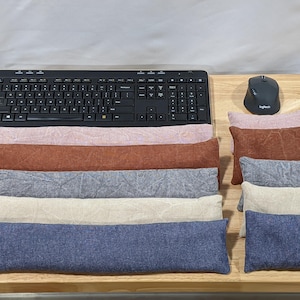 May include: A collection of rectangular fabric wrist rests and mouse pads in various colors including pink, rust, gray, cream, and blue. The items are arranged on a wooden surface alongside a black computer keyboard and a black computer mouse.