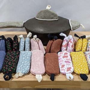 May include: A cast iron pot with a lid, featuring a grey fabric lid handle cover and two grey fabric side handle covers. In front, a collection of rectangular fabric pot holders in various colors and patterns, including floral, solid, and animal prints.