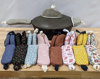 May include: A cast iron pot with a lid, featuring a grey fabric lid handle cover and two grey fabric side handle covers. In front, a collection of rectangular fabric pot holders in various colors and patterns, including floral, solid, and animal prints.