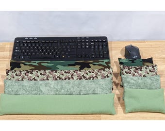 May include: A black computer keyboard and mouse positioned next to four long rectangular wrist rests and four smaller rectangular cushions. The items feature various green, brown, and black camouflage patterns and solid light green fabric textures.