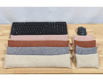 May include: A black computer keyboard and mouse accompanied by four long rectangular wrist rests and four smaller rectangular wrist rests in dusty pink, terracotta, slate blue, and beige, all featuring a subtle textured fabric pattern.