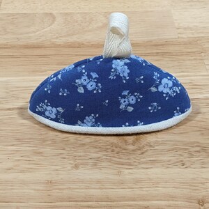 May include: A dome-shaped fabric tea cozy featuring a dark blue background with a light blue floral pattern. The item has a white fabric trim along the bottom edge and a small, off-white fabric loop attached to the top center for hanging.