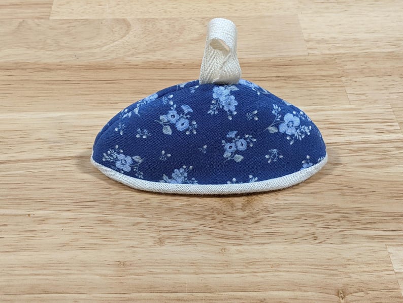 May include: A dome-shaped fabric tea cozy featuring a dark blue background with a light blue floral pattern. The item has a white fabric trim along the bottom edge and a small, off-white fabric loop attached to the top center for hanging.