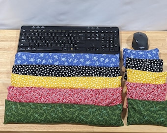 May include: A collection of rectangular fabric wrist rests and smaller rectangular fabric pouches featuring various patterns including blue floral, black paw prints, yellow floral, red floral, and green dragonfly designs, arranged near a black keyboard and mouse.
