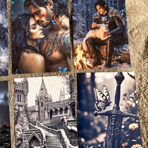 May include: A collection of fantasy art prints featuring romantic couples, a figure with a white fox by a fire, a castle, and a sword with a butterfly. The prints have a dark, moody aesthetic with detailed illustrations.