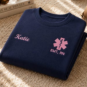 May include: A navy blue crewneck sweatshirt featuring light pink embroidery on the front. The embroidery includes the name Katie in script font, a Star of Life medical symbol, and the text EMT, RN positioned below the medical emblem.