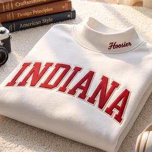 May include: A white mock neck sweatshirt featuring the word INDIANA in large red block letters across the chest and the word Hoosier embroidered in red on the collar. The garment is made of a soft fabric and has a ribbed neckline.
