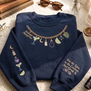 May include: A navy blue crewneck sweatshirt featuring gold chain embroidery with charms including a margarita, crescent moon, candle, potion bottle, lime slice, and palm leaf. The text reads Midnight Margaritas and Put the Time in the Coconut And Drink It All Up.