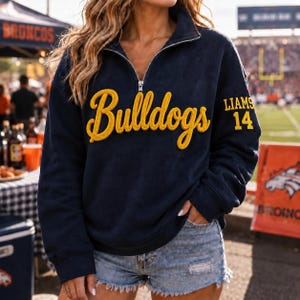 May include: A navy blue quarter-zip pullover sweatshirt featuring the word Bulldogs in large, yellow, textured script lettering across the chest. The left sleeve displays the text LIAMS and the number 14 in yellow block lettering.