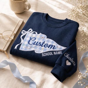 May include: A navy blue crewneck sweatshirt featuring an embroidered light blue and white gingham pennant design on the chest. The pennant includes the text Custom and SCHOOL NAME. The sleeve is embroidered with the name Charlotte and a heart.