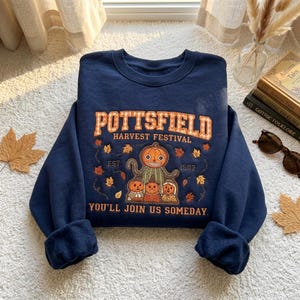 Pode incluir: Uma camisola azul-marinho de gola redonda com uma impressão gráfica com o texto POTTSFIELD HARVEST FESTIVAL EST 1502 YOU'LL JOIN US SOMEDAY. O design inclui uma grande personagem de abóbora central, figuras de abóbora mais pequenas e folhas.